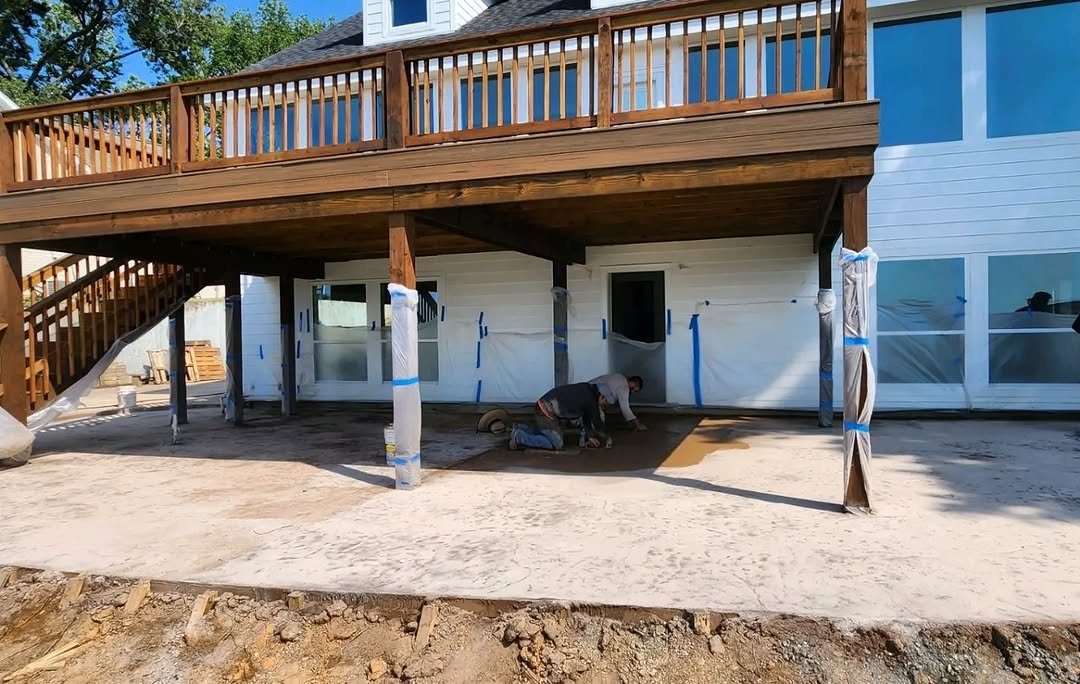 Willmer Construction's crew finishing a new concrete patio under the wraparound deck of a two-story white home, with the posts and windows wrapped in plastic to protect them.