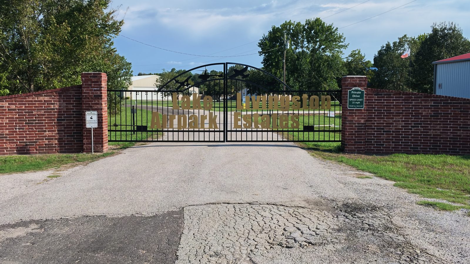 Closed metal entrance gate lettered Lake Livingston Airpark Estates, brick pillars either side, aircraft hangars visible beyond.