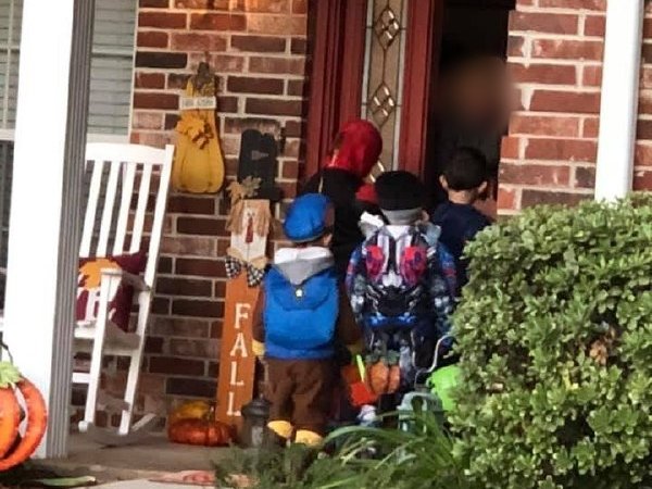 Kids in costume, including a blue cap and backpack and a superhero, at a brick front porch decorated for fall with pumpkins and a wooden FALL sign.