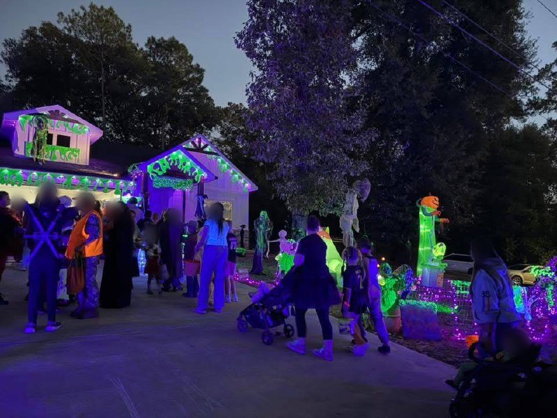 Families in costume crowd a street at night outside a house lit in purple and green, with glowing inflatable monsters and skeletons in the yard.